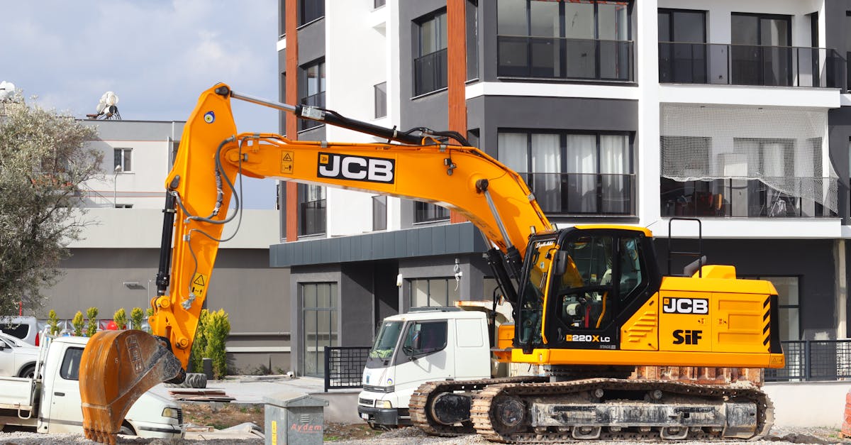 A yellow excavator operating at a city construction site in front of modern buildings.